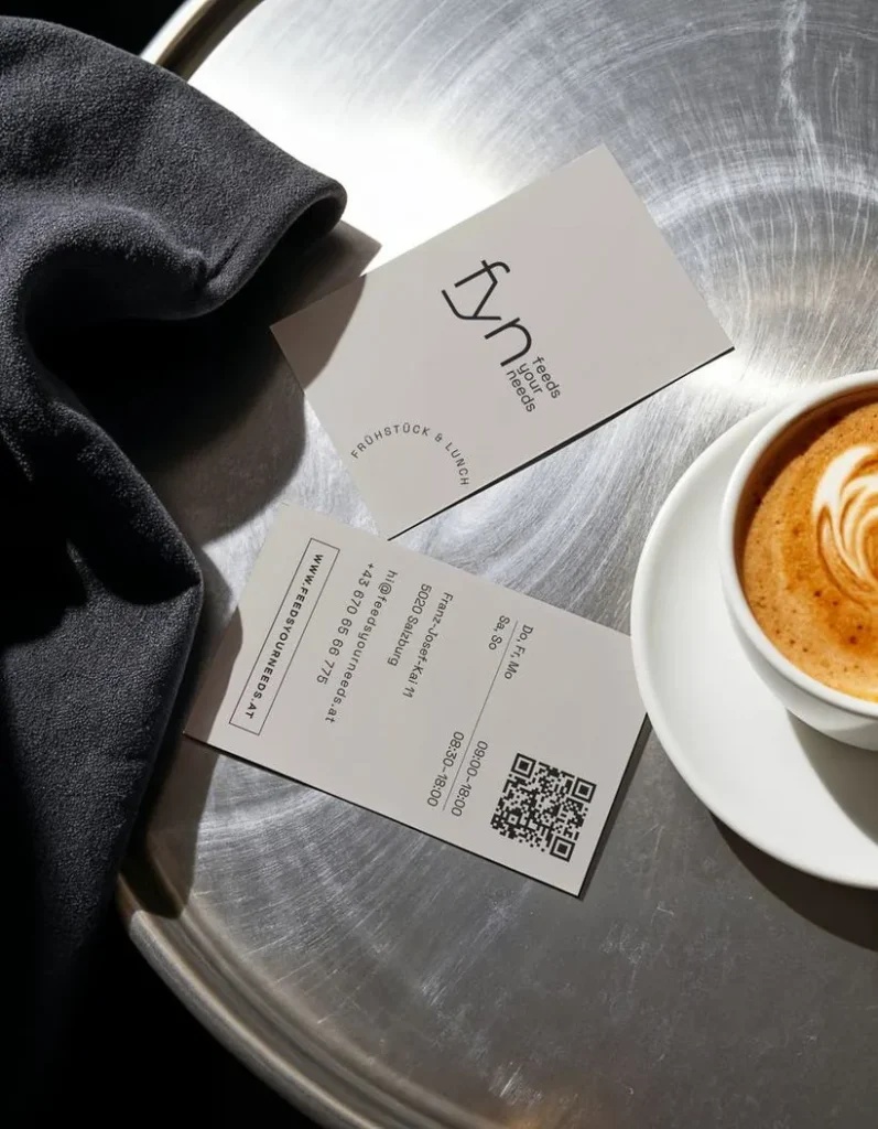 Auf einem runden Metalltisch liegen zwei Visitenkarten des Restaurants „Fyn“ neben einer weißen Tasse Cappuccino und einem dunklen Tuch. Auf der einen Karte sind die Kontaktdaten und ein QR-Code zu sehen, auf der anderen das Logo des Restaurants.